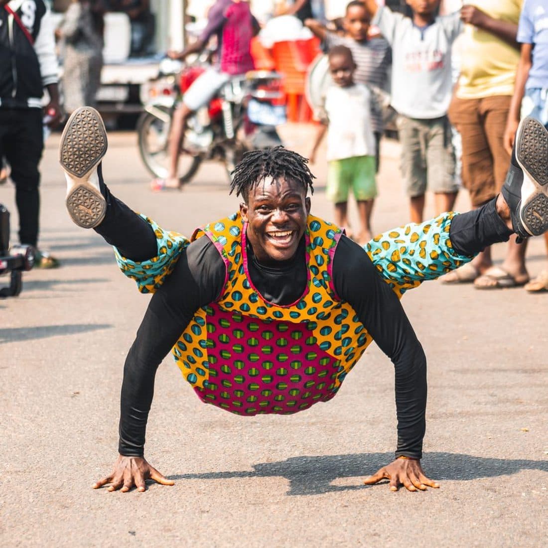 An African acrobat in printed costume holding a balance on his hands before a street crowd