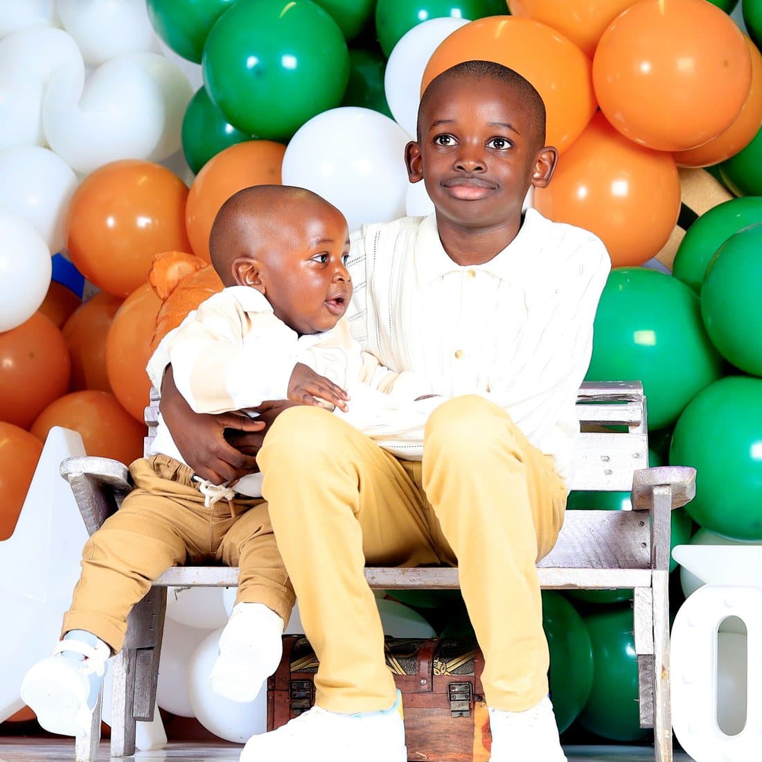 Two young brothers seated before a green and orange balloon garland at a birthday celebration