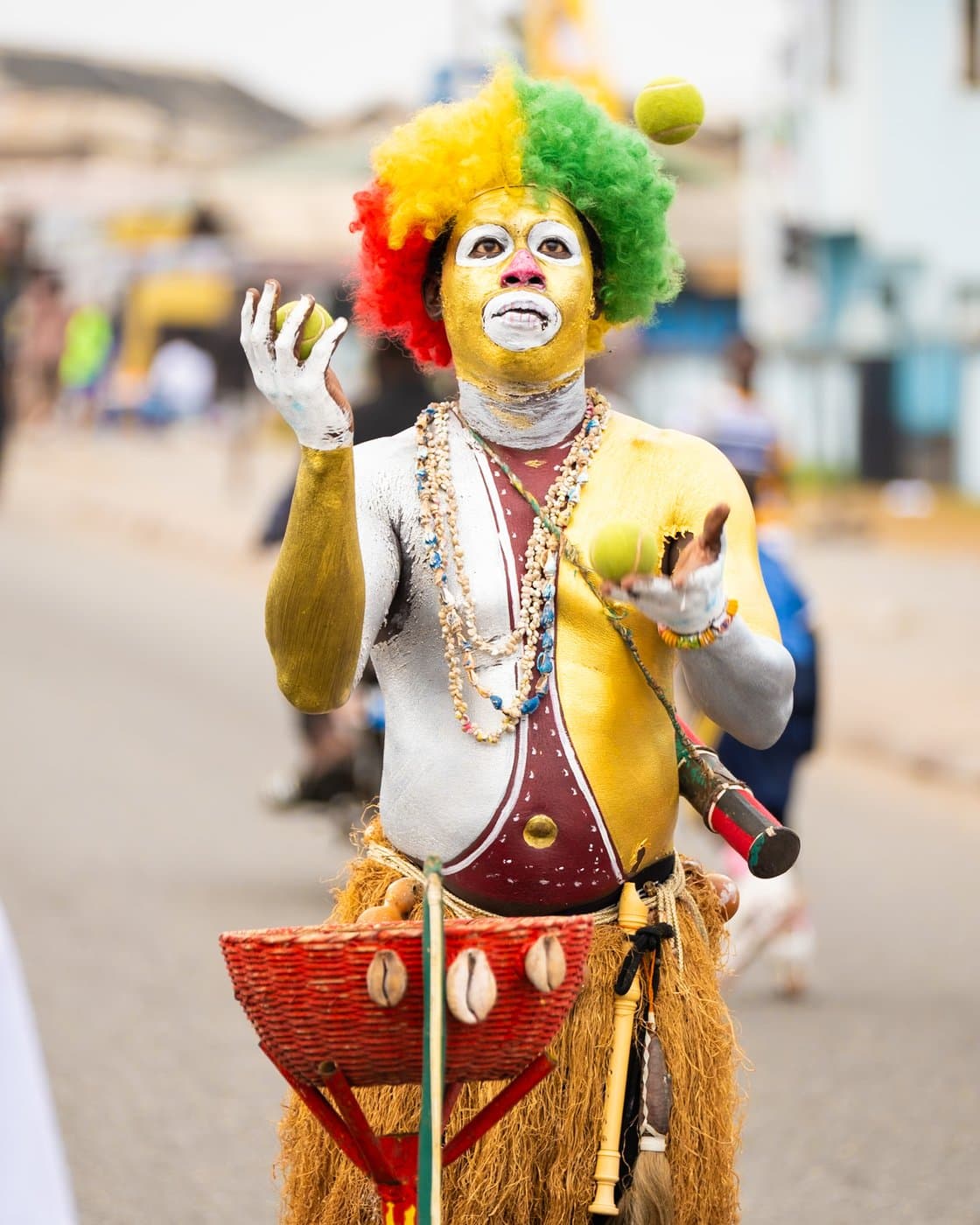 A West African street clown in full regalia — rainbow wig, painted face, cowrie shells and raffia — juggling at a street festival